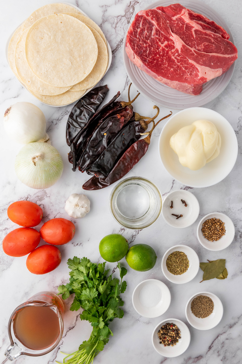 Essential ingredients for homemade quesabirria tacos including chuck roast, dried guajillo chilies, tortillas, onions, garlic, tomatoes, spices, cilantro, and lime.