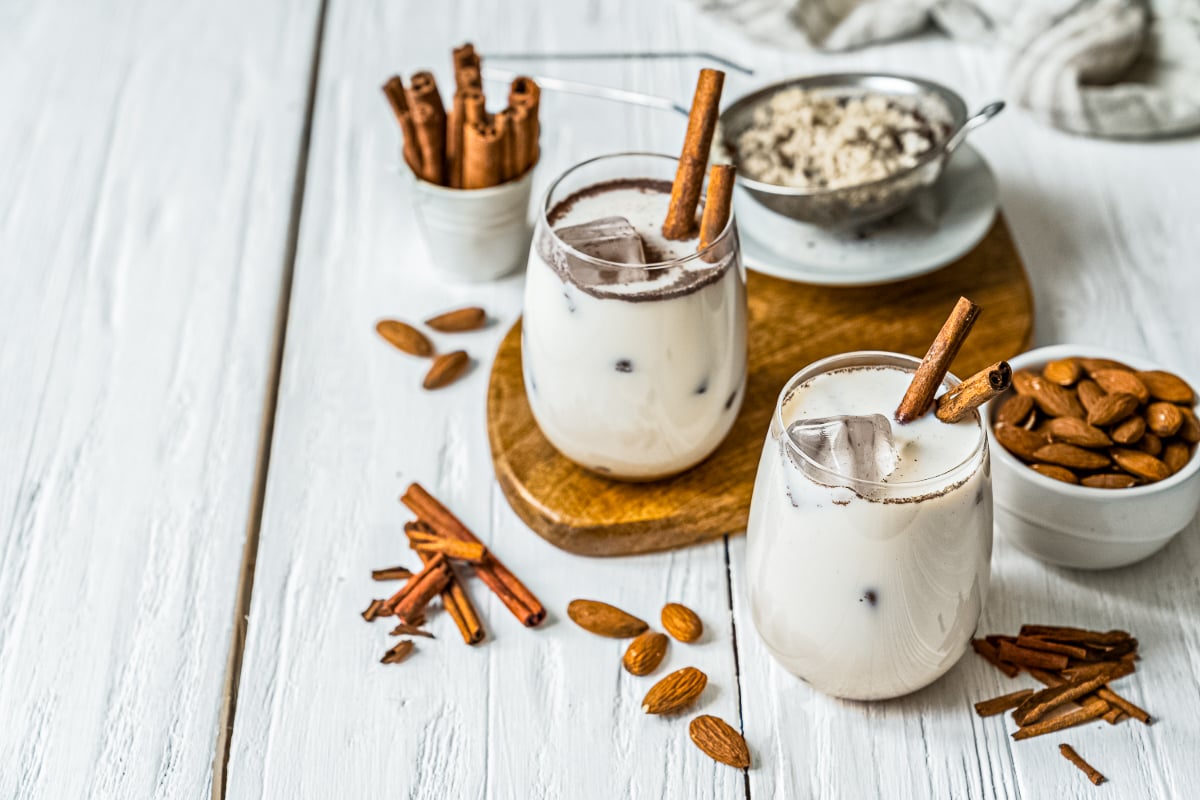 Two glasses of creamy iced Mexican horchata garnished with cinnamon sticks, surrounded by almonds and cinnamon on a white wooden table.