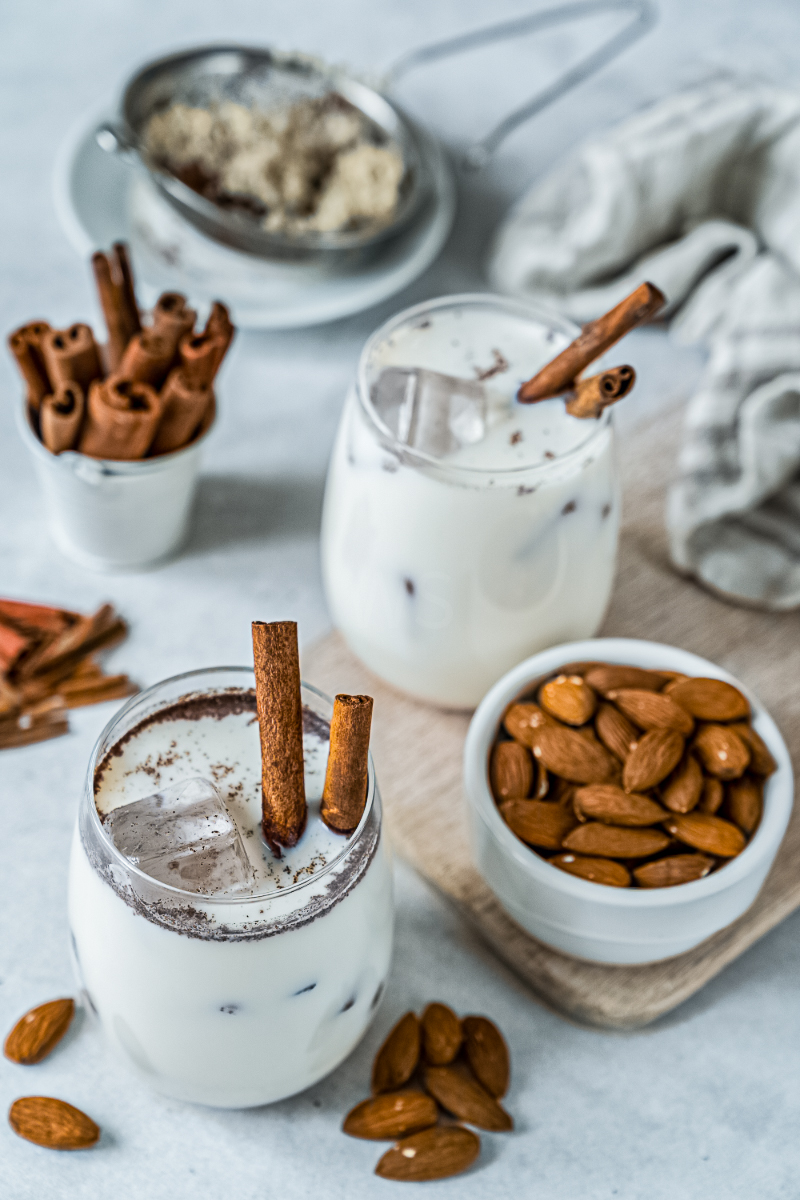 Creamy iced Mexican horchata in two glasses with cinnamon stick garnish, served beside a bowl of almonds and extra cinnamon on a light tabletop.