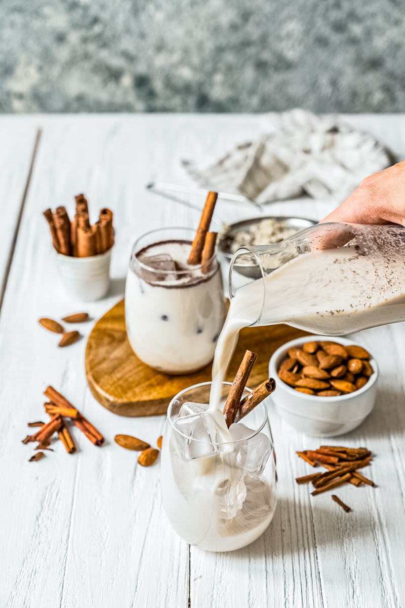 Homemade Mexican horchata being poured over ice into a glass, with cinnamon sticks, almonds, and extra chilled glasses on a white wooden surface.