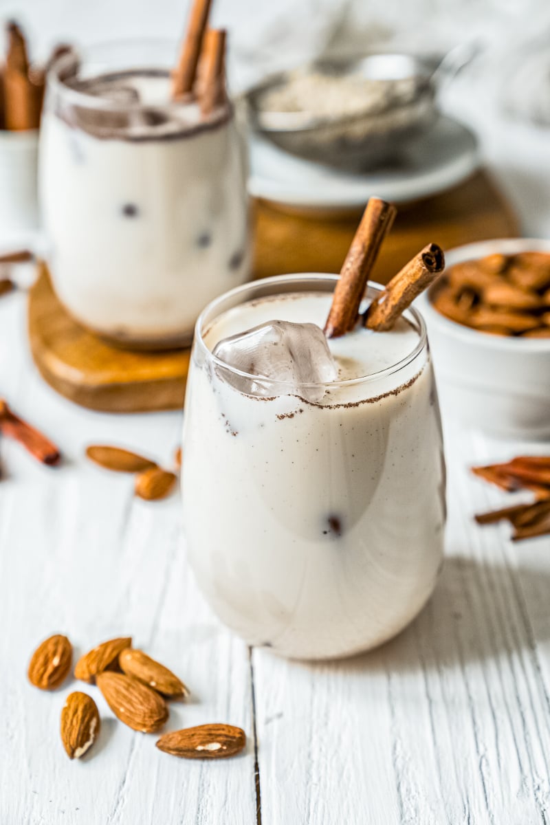 Creamy homemade Mexican horchata served over ice in stemless glasses with cinnamon stick garnish, scattered almonds, and a rustic kitchen backdrop.
