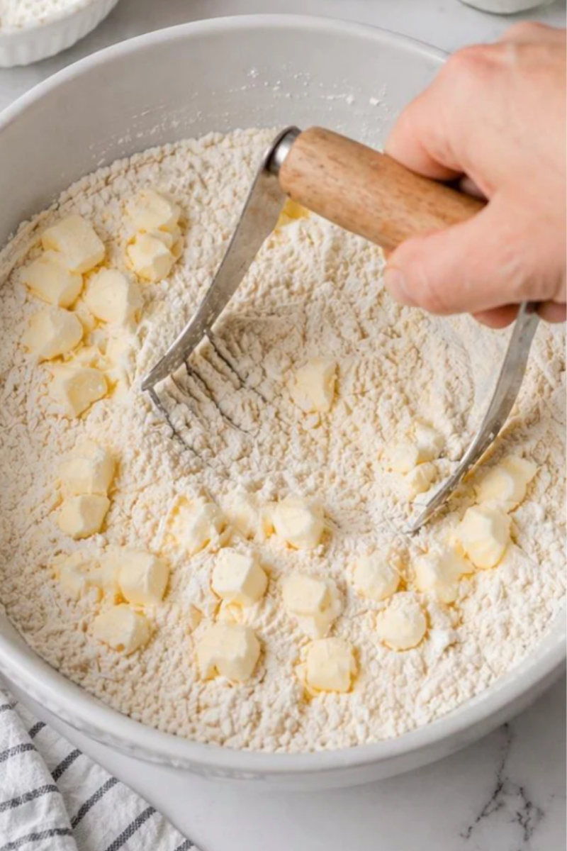 Cold butter being cut into the flour mixture with a pastry cutter for flaky strawberry shortcake biscuits.