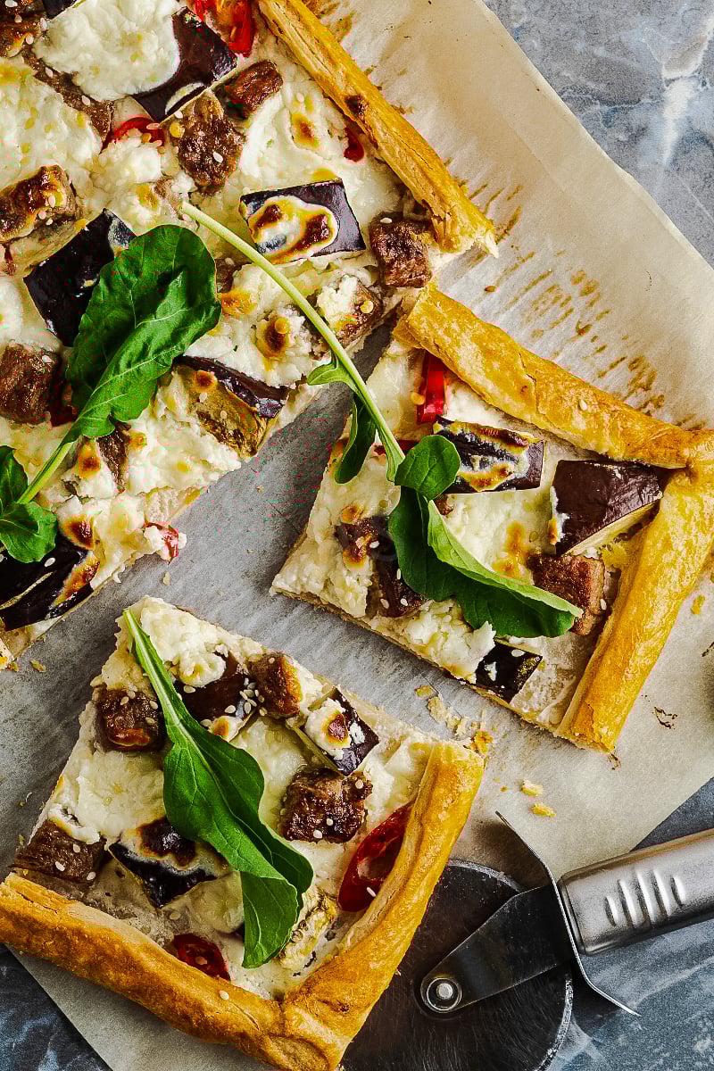 Golden puff pastry tart topped with roasted eggplant, cherry tomatoes, fresh herbs, and arugula on a rustic baking sheet.