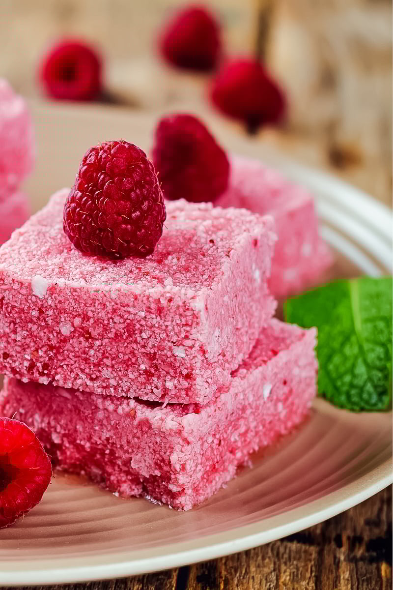 Close-up of creamy raspberry coconut butter fudge squares on a white plate, topped with fresh raspberries and a dark chocolate drizzle, showcasing a vibrant pink texture.