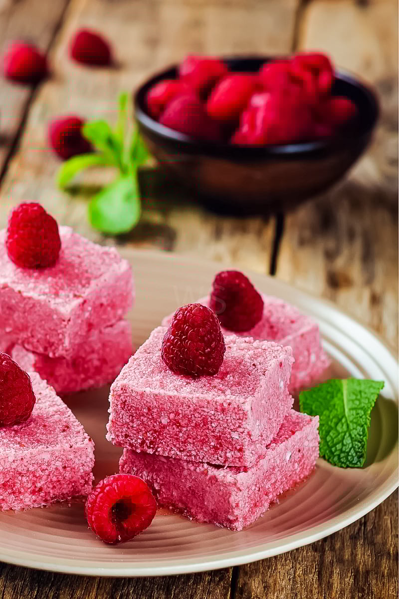 Close-up of creamy raspberry coconut butter fudge squares on a white plate, topped with fresh raspberries and a dark chocolate drizzle, showcasing a vibrant pink texture.