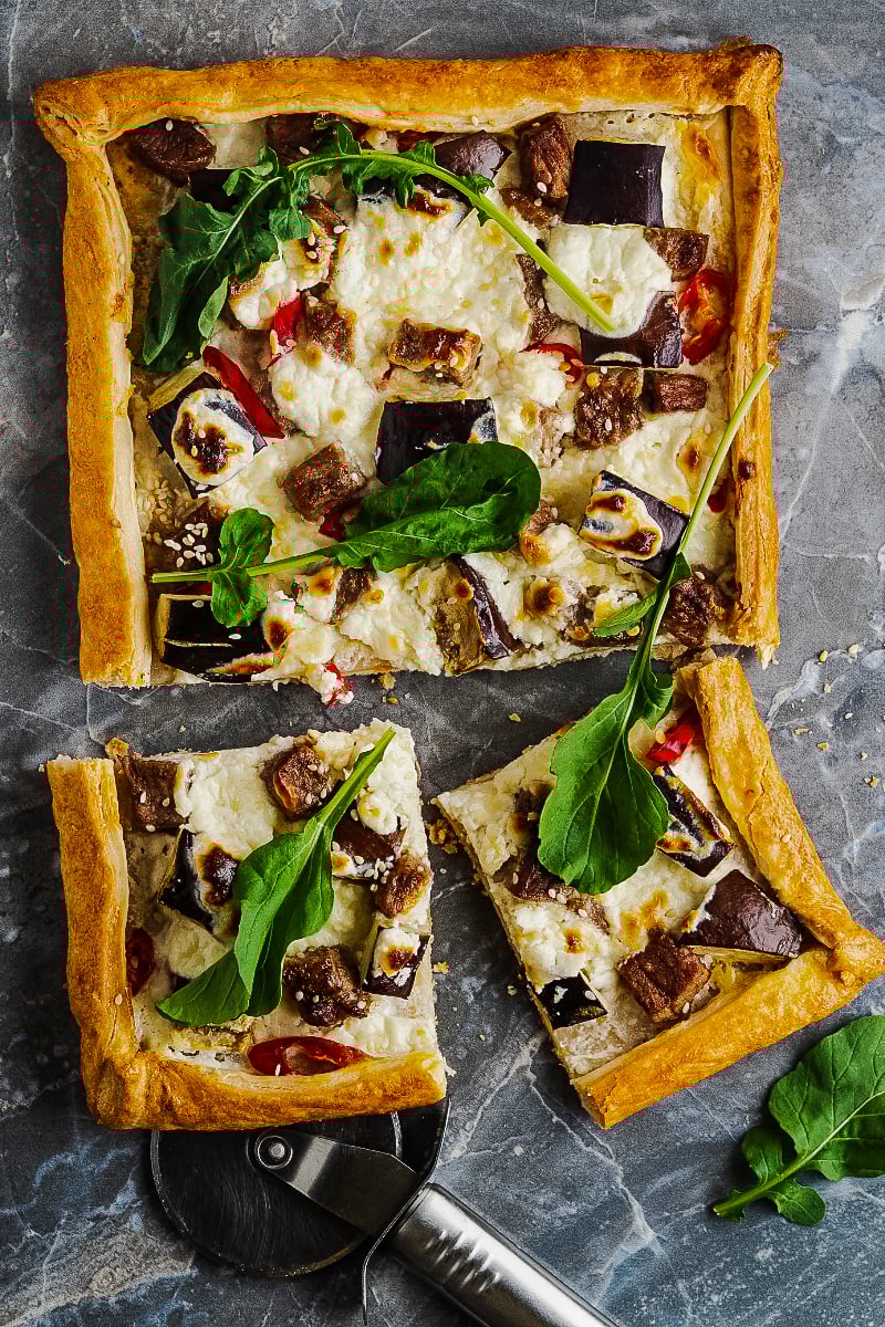 Golden puff pastry tart topped with roasted eggplant, cherry tomatoes, fresh herbs, and arugula on a rustic baking sheet.