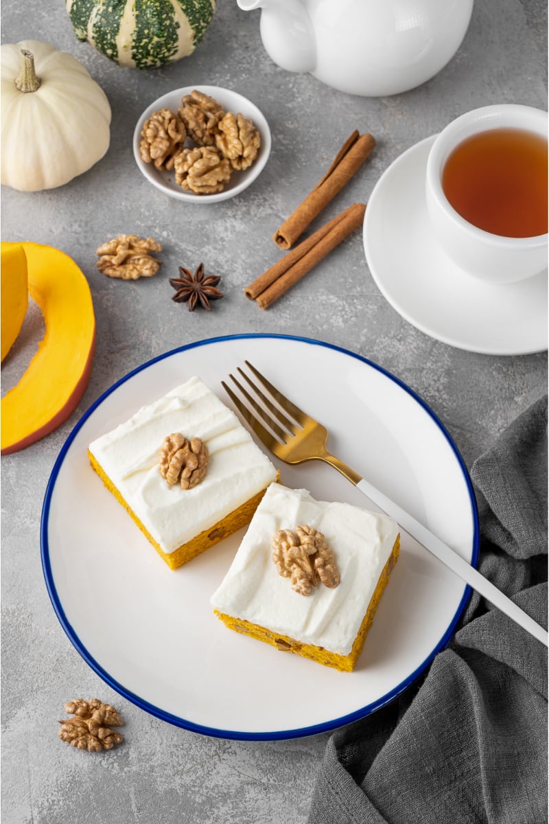 Slice of spiced pumpkin cake topped with creamy walnut-studded frosting and cinnamon dusting on a rustic fall plate.