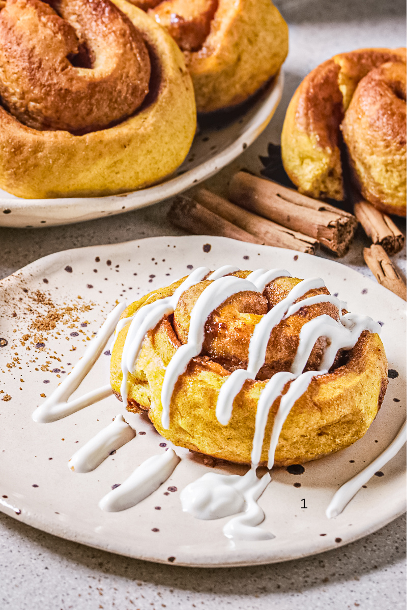 Freshly baked pumpkin cinnamon buns with swirls of cinnamon sugar, drizzled with creamy vanilla glaze, served on a rustic plate for cozy autumn breakfast.