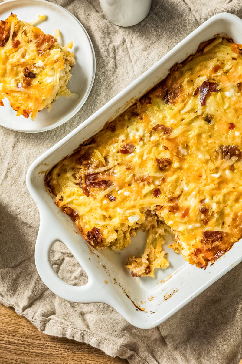 Amish breakfast casserole with bacon, eggs, hash browns, and melted cheddar cheese baked until golden in a white casserole dish with a slice served on a plate.
