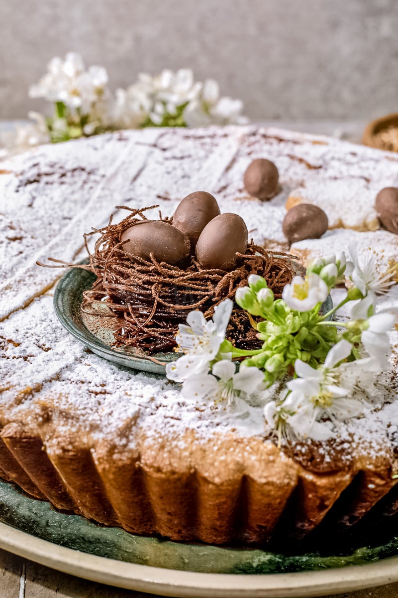 Pastiera Napoletana Italian Easter tart dusted with powdered sugar, topped with a chocolate nest and mini eggs, styled with fresh spring blossoms for a festive holiday dessert presentation.