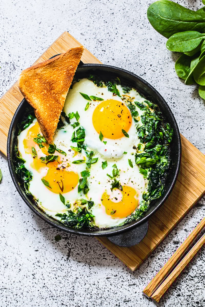 Green shakshuka with spinach and eggs cooked in a cast iron skillet, topped with fresh herbs and served with toasted bread.