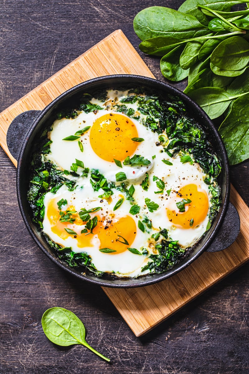 Green shakshuka with spinach and eggs cooked in a cast iron skillet, garnished with fresh herbs and served on a rustic wooden board.