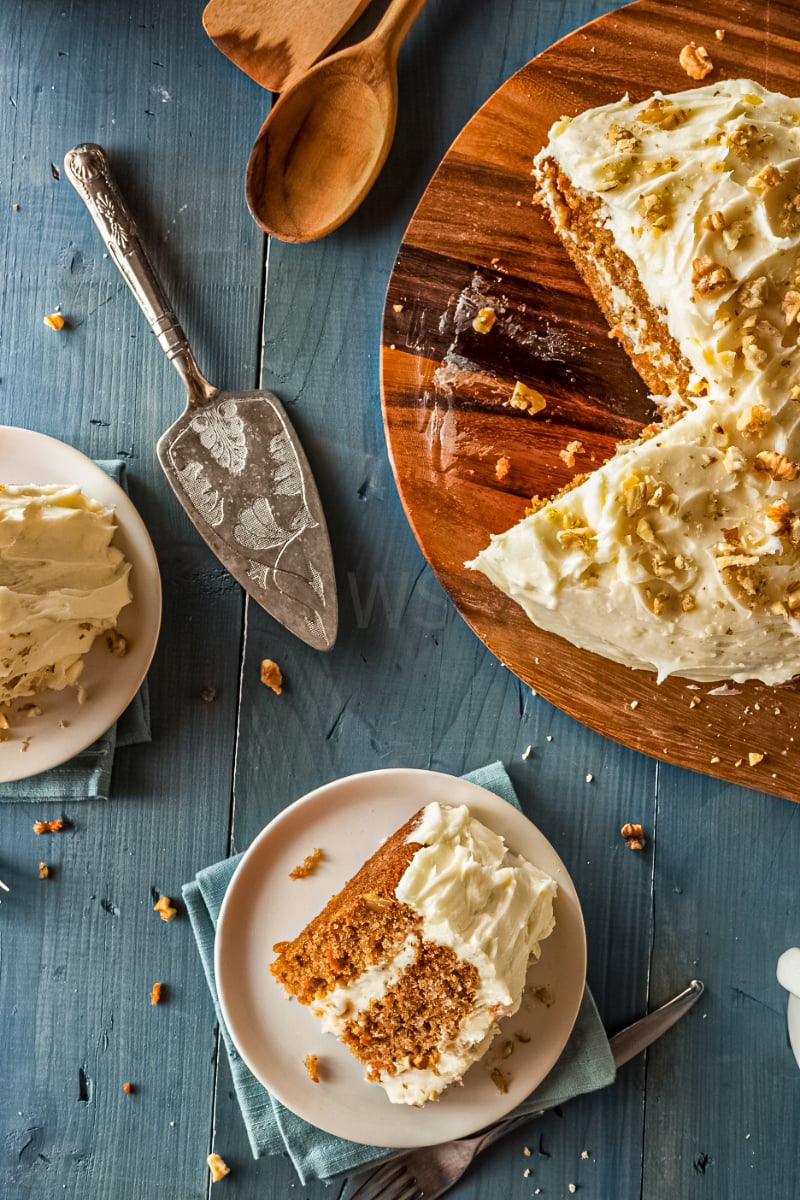 Healthy homemade carrot cake with cream cheese frosting and chopped walnuts on a wooden serving board, with a slice plated on a blue rustic table
