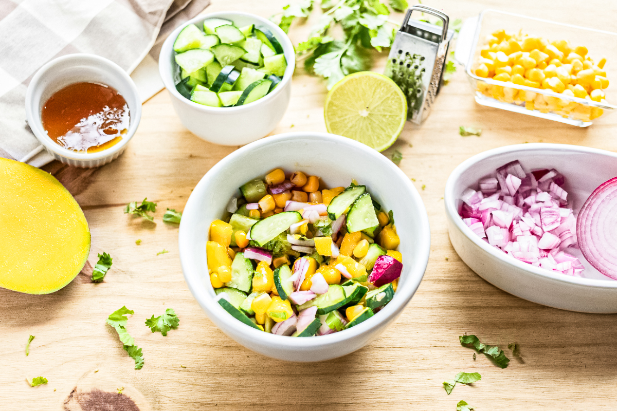 ngredients for cucumber mango salsa including diced mango, cucumber, red onion, corn, lime, cilantro, and honey on a wooden surface.