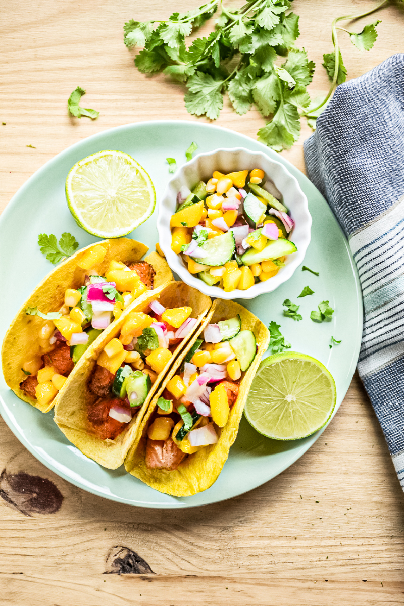 Pan-seared salmon tacos topped with cucumber mango salsa served on corn tortillas with lime wedges and fresh cilantro.