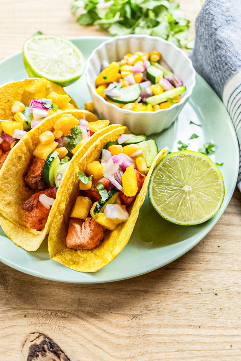 Salmon tacos with cucumber mango salsa served on corn tortillas with fresh lime wedges and a bowl of salsa on a plate.