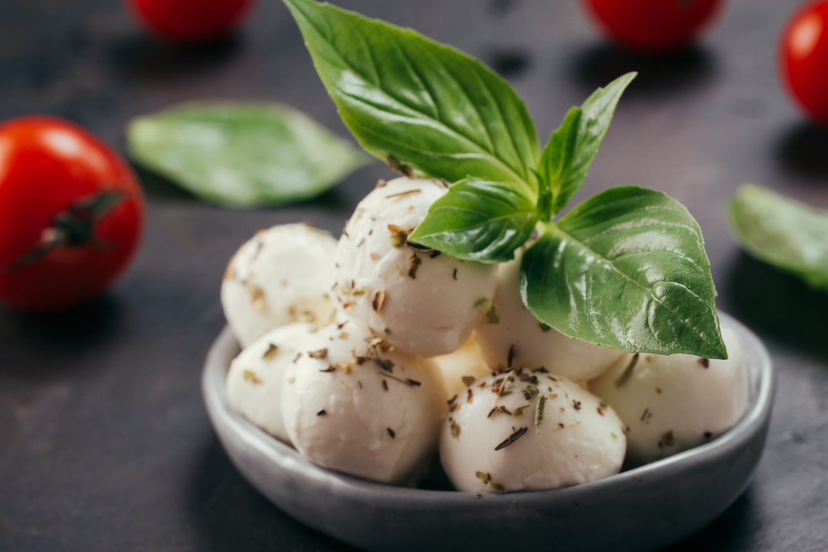 Fresh mozzarella pearls topped with basil leaves and herbs with cherry tomatoes in the background, classic caprese ingredients.