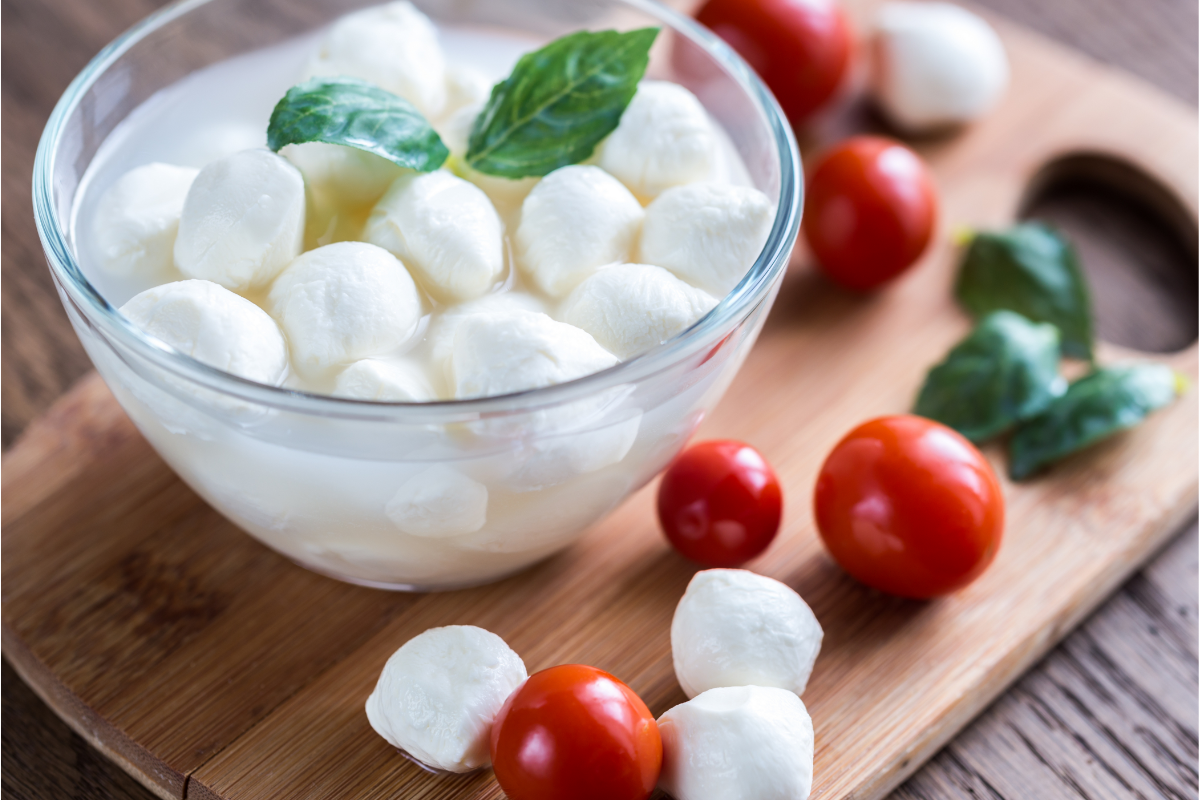 Fresh mozzarella pearls in a bowl with cherry tomatoes and basil on a wooden board, ingredients for caprese avocado recipe.