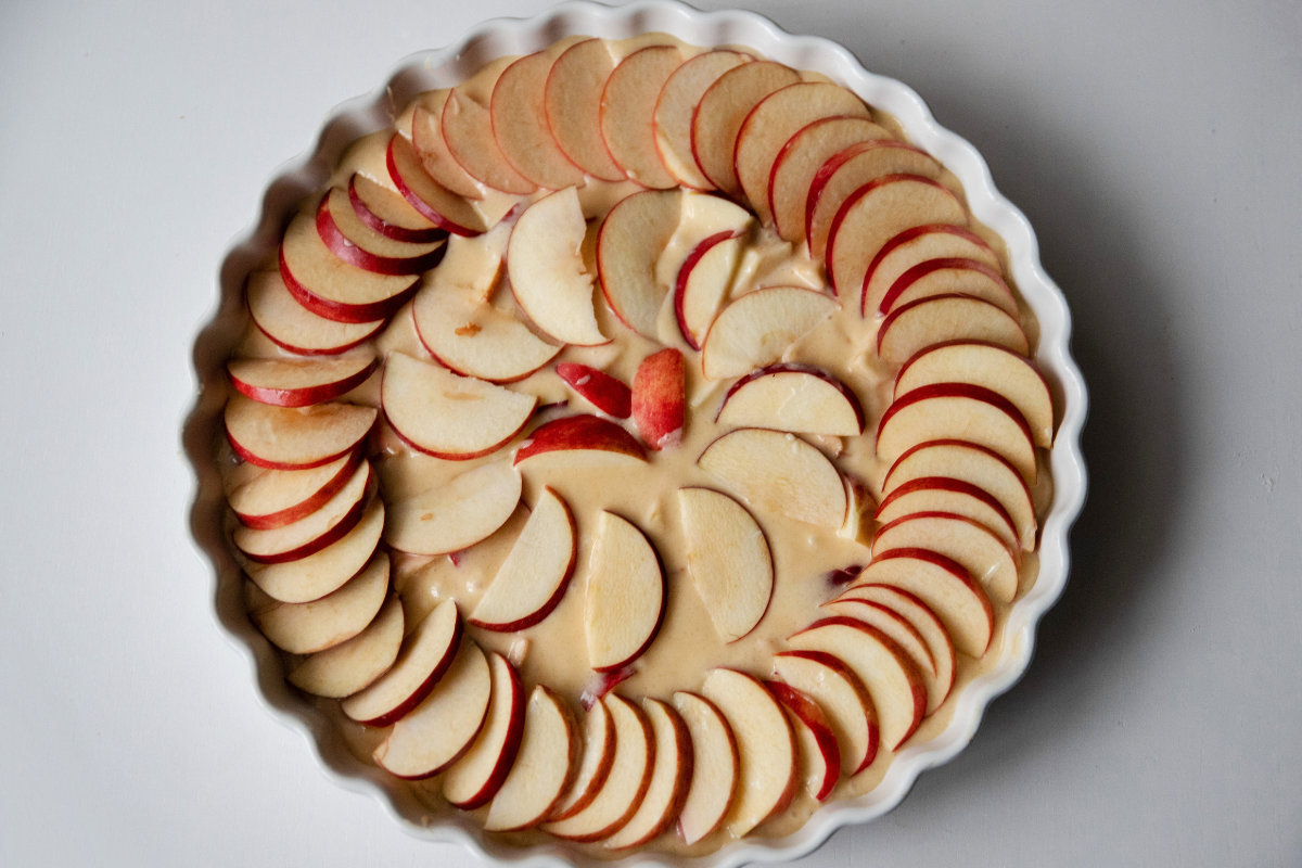 Thinly sliced red apples arranged in a circular pattern over batter in a white baking dish, ready to bake.