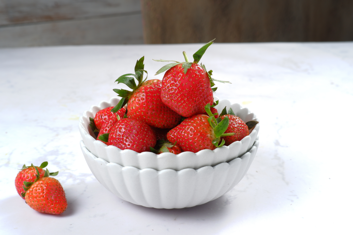 Fresh strawberries in a white ceramic bowl on a light marble surface with one strawberry beside the bowl.
