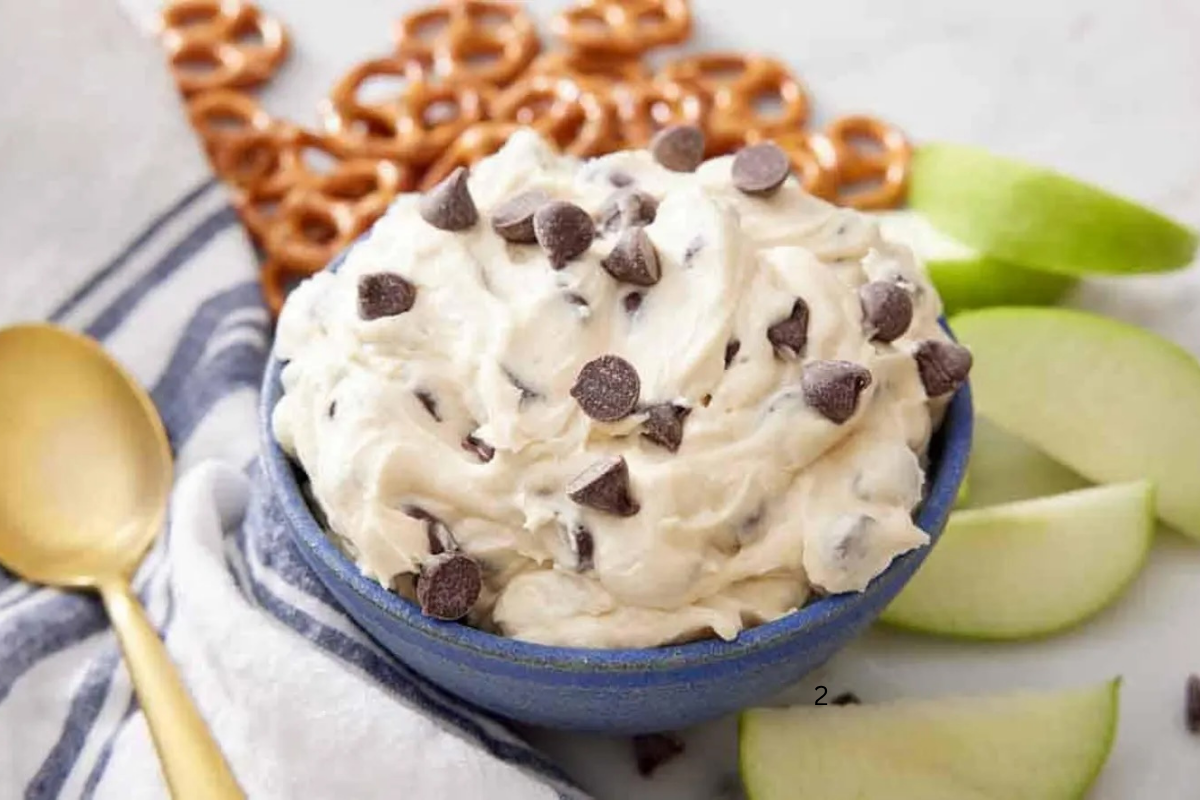 Close-up of creamy cookie dough dip topped with chocolate chips in a blue bowl, served with pretzels and sliced green apples.