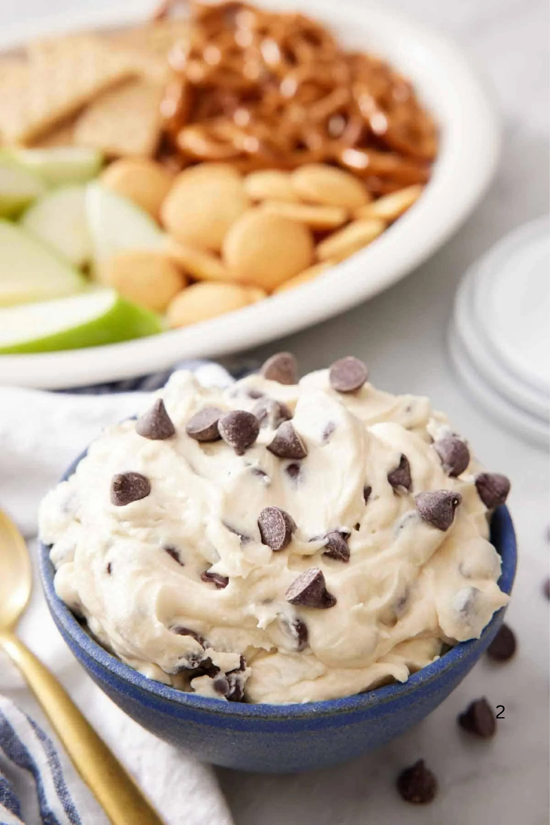 Close-up of creamy cookie dough dip with chocolate chips in a blue bowl, with pretzels, cookies, and sliced green apples in the background.
