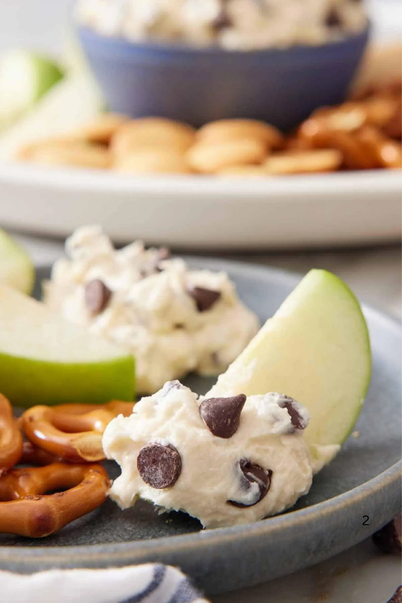 Apple slice dipped in creamy cookie dough dip with chocolate chips, served with pretzels on a plate.