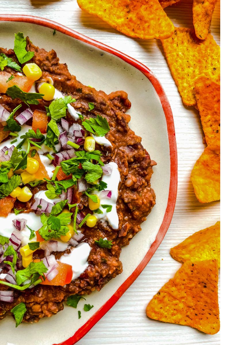 Close-up of black bean dip topped with red onion, corn, tomato, cilantro, and a creamy drizzle, served with tortilla chips on the side.