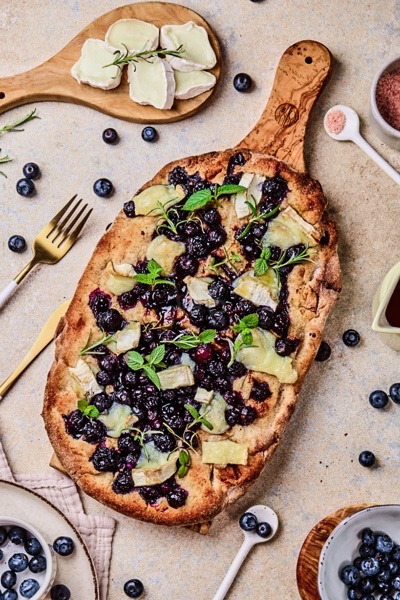 Overhead view of blueberry goat cheese flatbread with baked blueberries, melted goat cheese, fresh mint, rosemary, and crisp golden crust served on a wooden board.