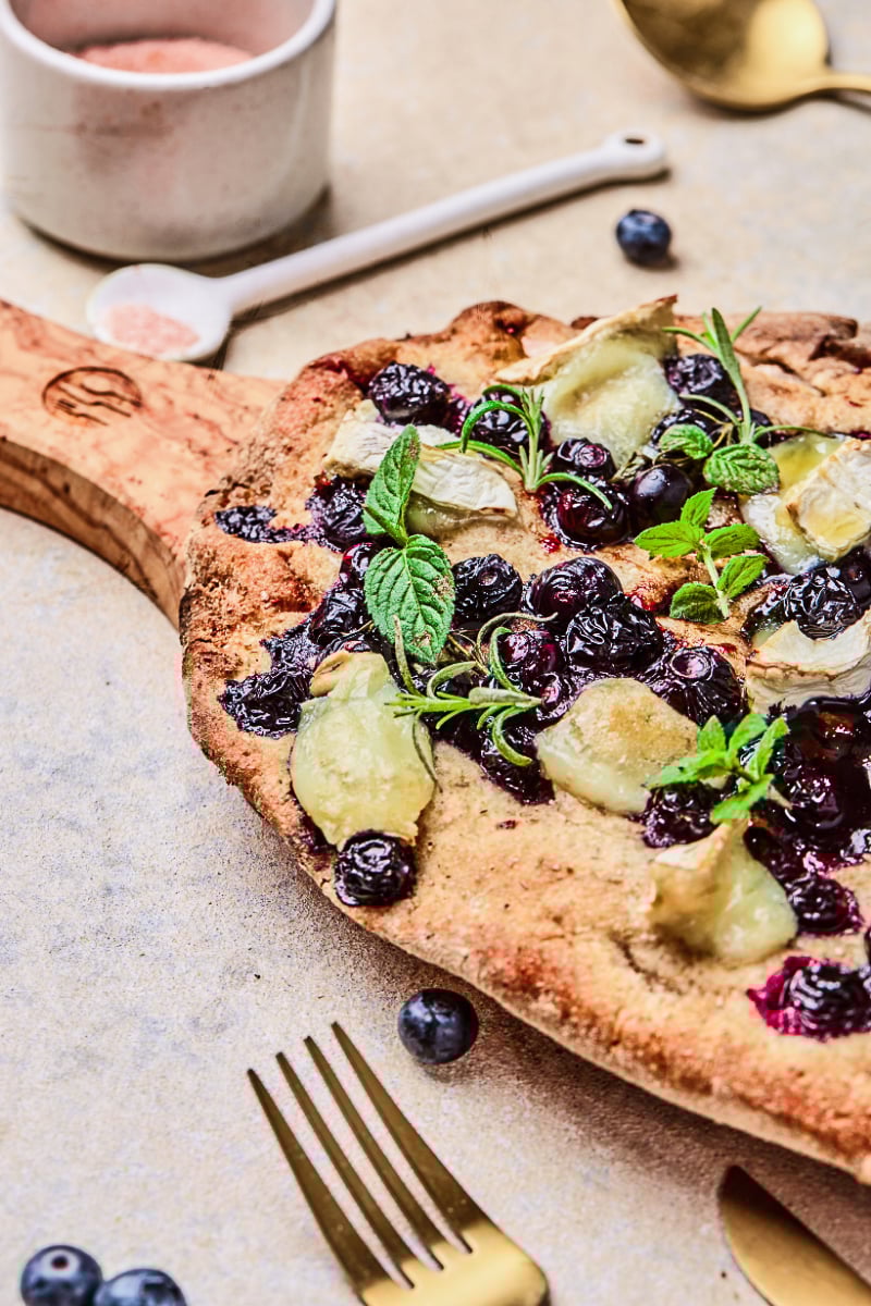 Blueberry goat cheese flatbread with baked blueberries, melted goat cheese, fresh mint, rosemary, and a crisp golden crust served on a wooden board as an easy appetizer.