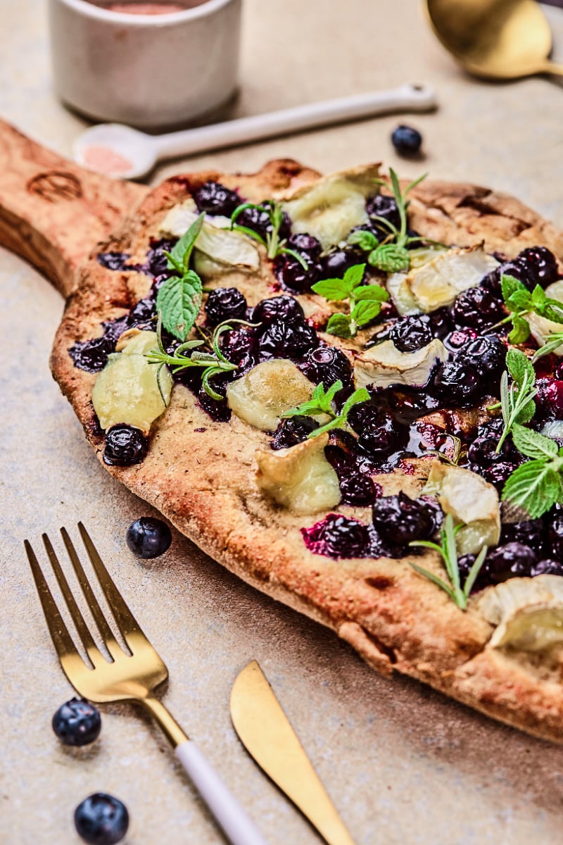 Blueberry goat cheese flatbread on a wooden board with baked blueberries, melted goat cheese, fresh mint, rosemary, and a crisp golden crust for a sweet-savory appetizer.
