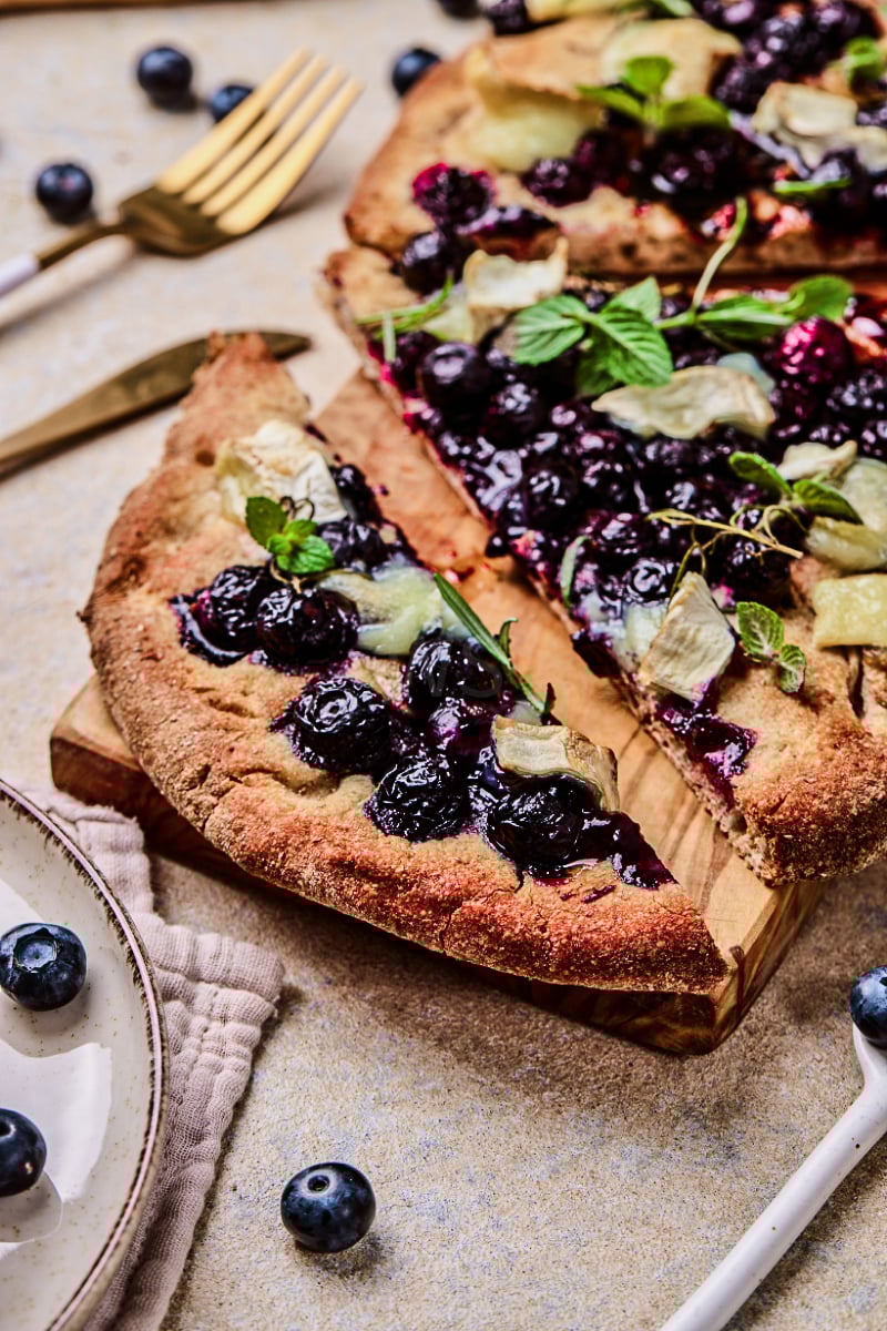 Blueberry goat cheese flatbread with a crisp golden crust, juicy baked blueberries, creamy melted goat cheese, fresh mint, and sliced pieces served on a wooden board.