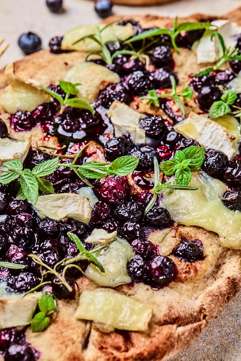 Blueberry goat cheese flatbread topped with glossy baked blueberries, creamy melted goat cheese, fresh mint, rosemary, and a crisp golden crust for an easy appetizer.