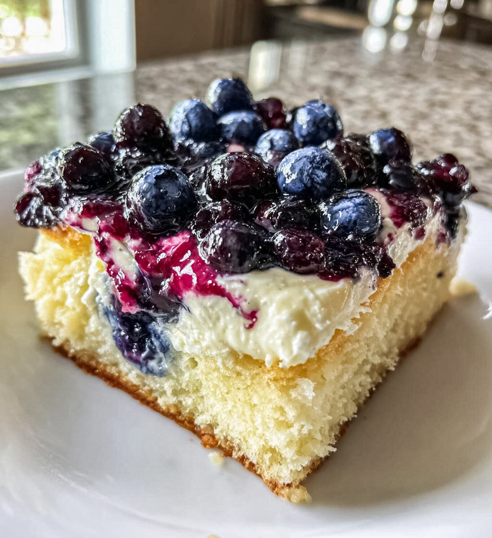 Blueberry lemon cheesecake square on a plate, topped with glossy blueberries, showing creamy cheesecake layer, fluffy cake base, and juicy berry topping.