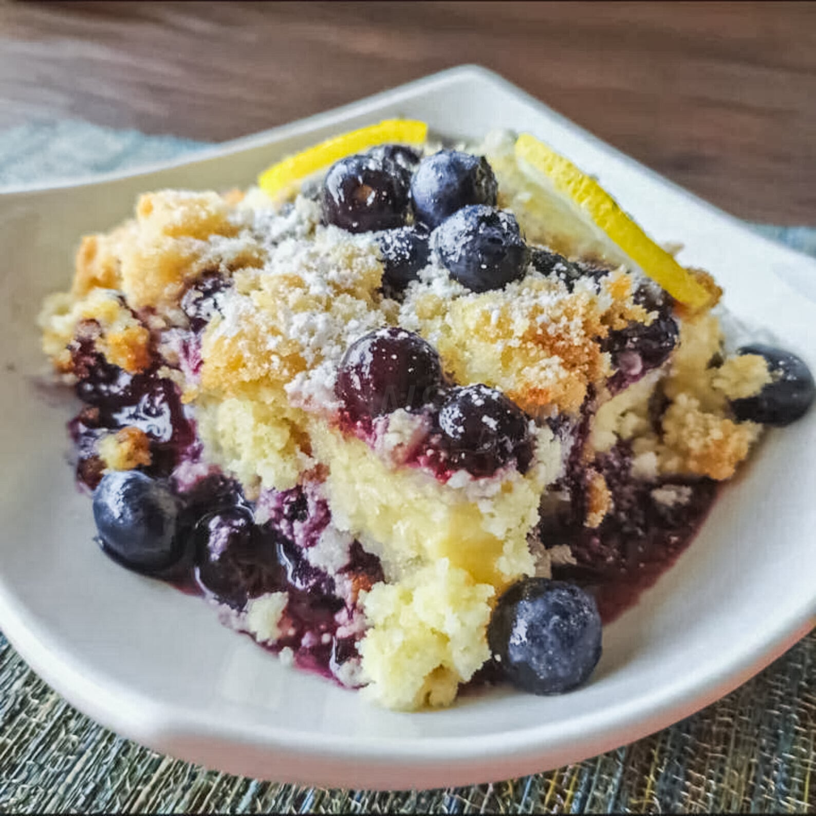 Blueberry lemon cheesecake dump cake on a plate, topped with fresh blueberries and powdered sugar, showing buttery cake, creamy filling, and juicy berry layers