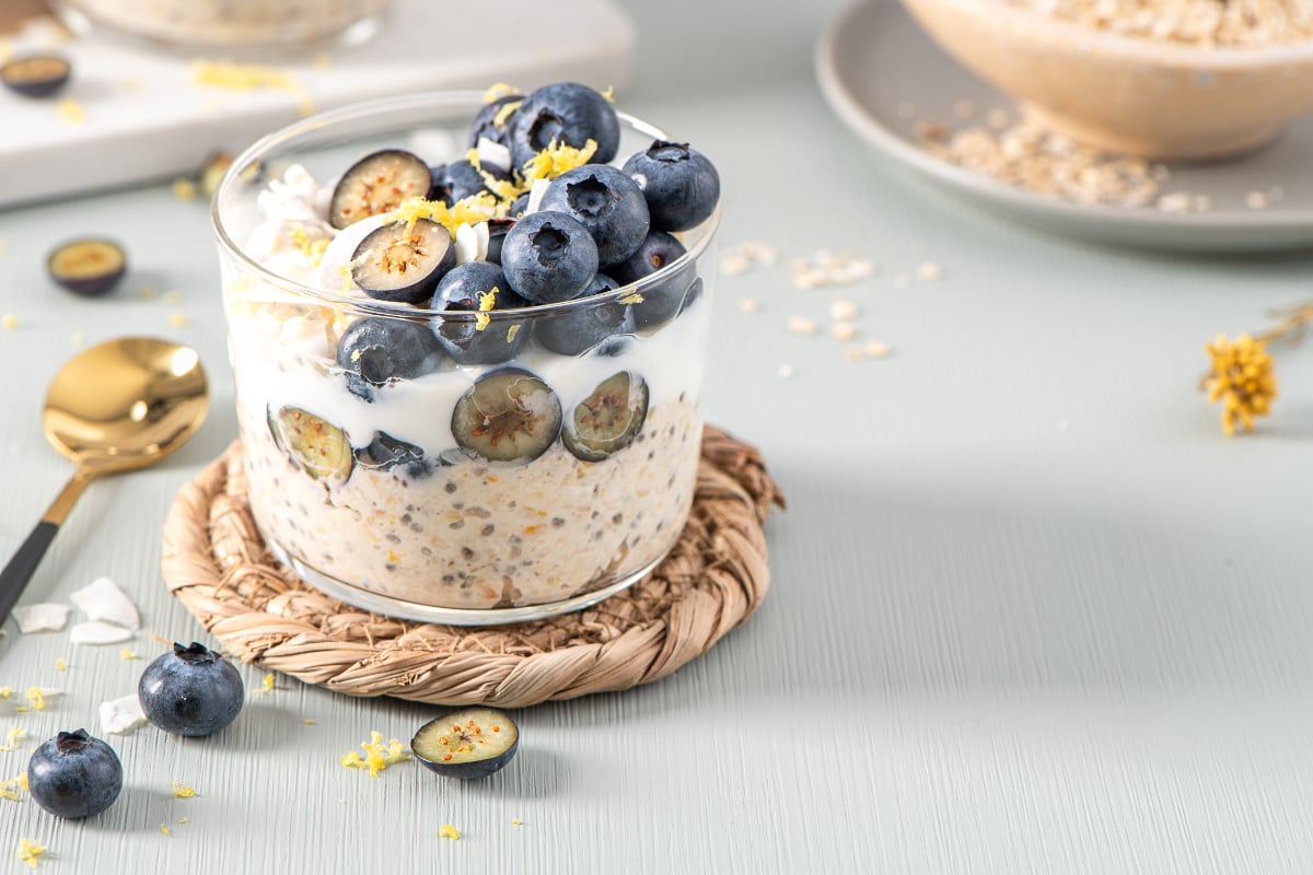 Creamy blueberry lemon overnight oats in a glass bowl topped with whole and sliced blueberries, lemon zest, and coconut flakes on a woven coaster.