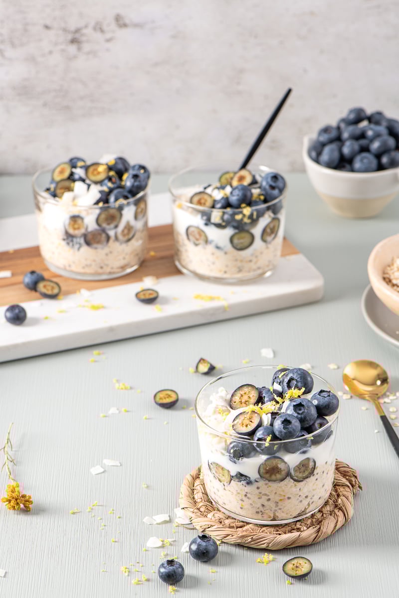 Blueberry lemon overnight oats in clear glass cups topped with whole and sliced blueberries, lemon zest, and coconut flakes, styled with a gold spoon and extra blueberries.