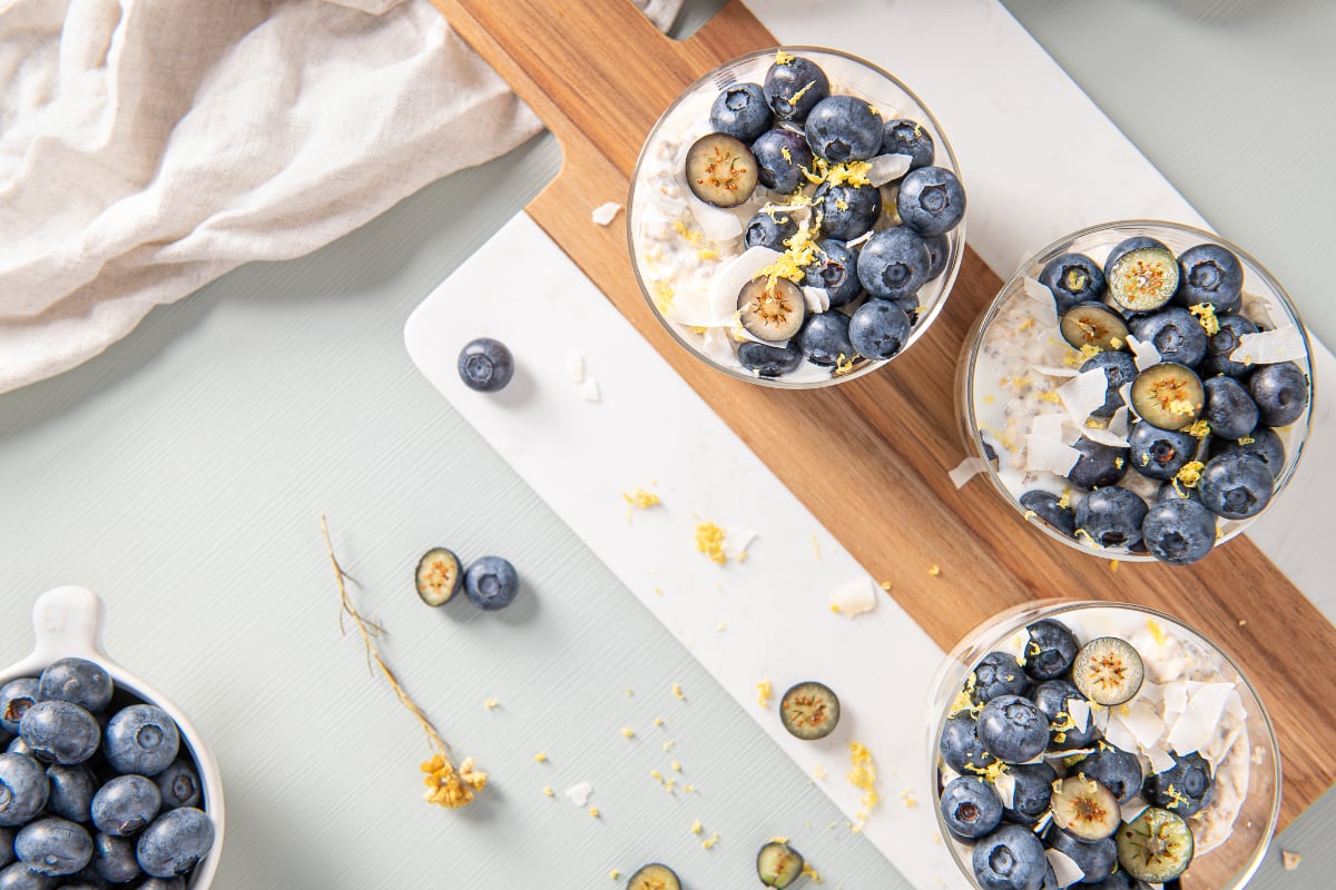 Three cups of blueberry lemon overnight oats topped with whole and sliced blueberries, lemon zest, and coconut flakes on a marble and wood serving board.