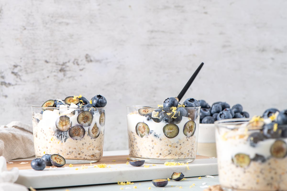 Three glass cups of blueberry lemon overnight oats layered with yogurt and topped with whole and sliced blueberries, lemon zest, and coconut flakes.