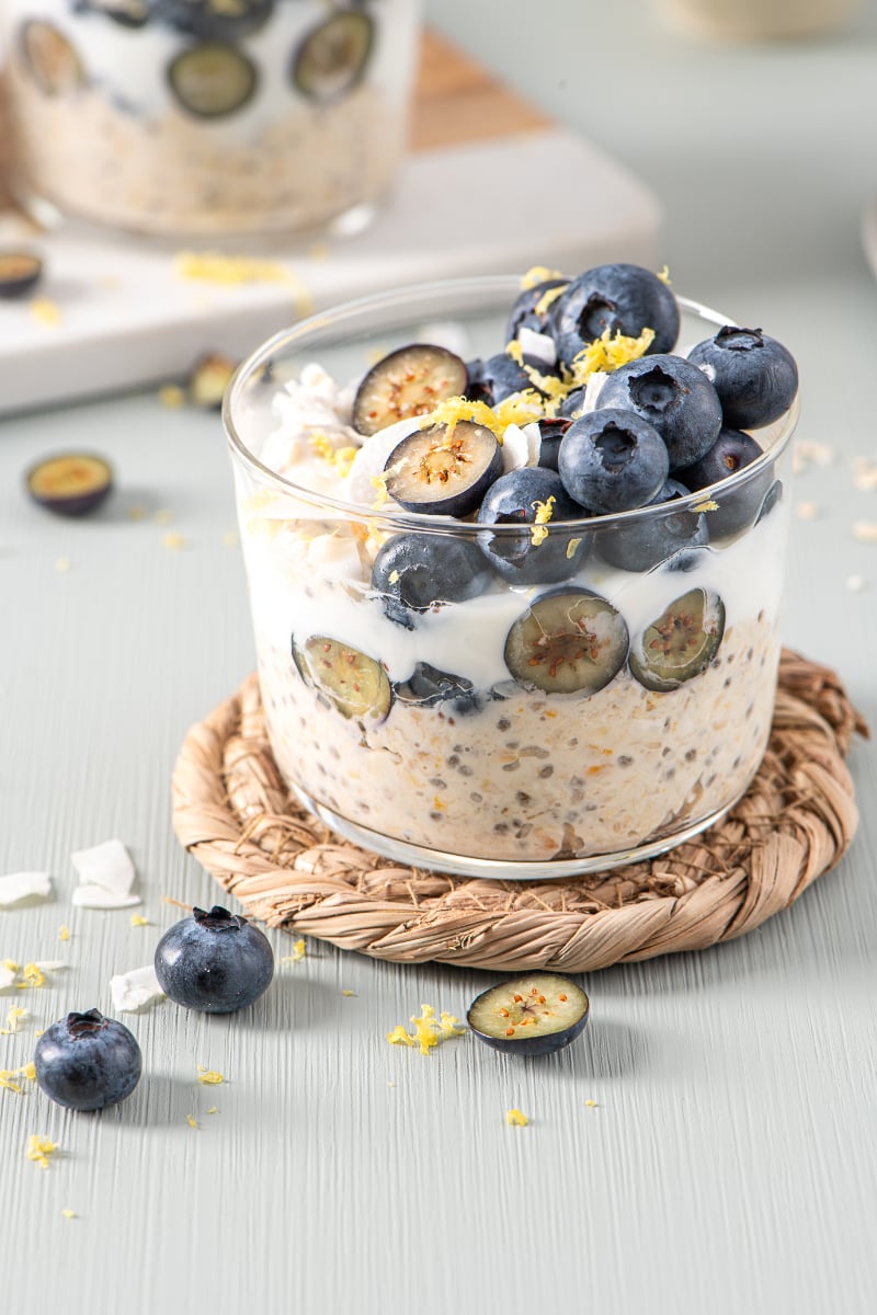 Creamy blueberry lemon overnight oats in a clear glass bowl, topped with whole and sliced blueberries, lemon zest, and coconut flakes on a woven coaster.