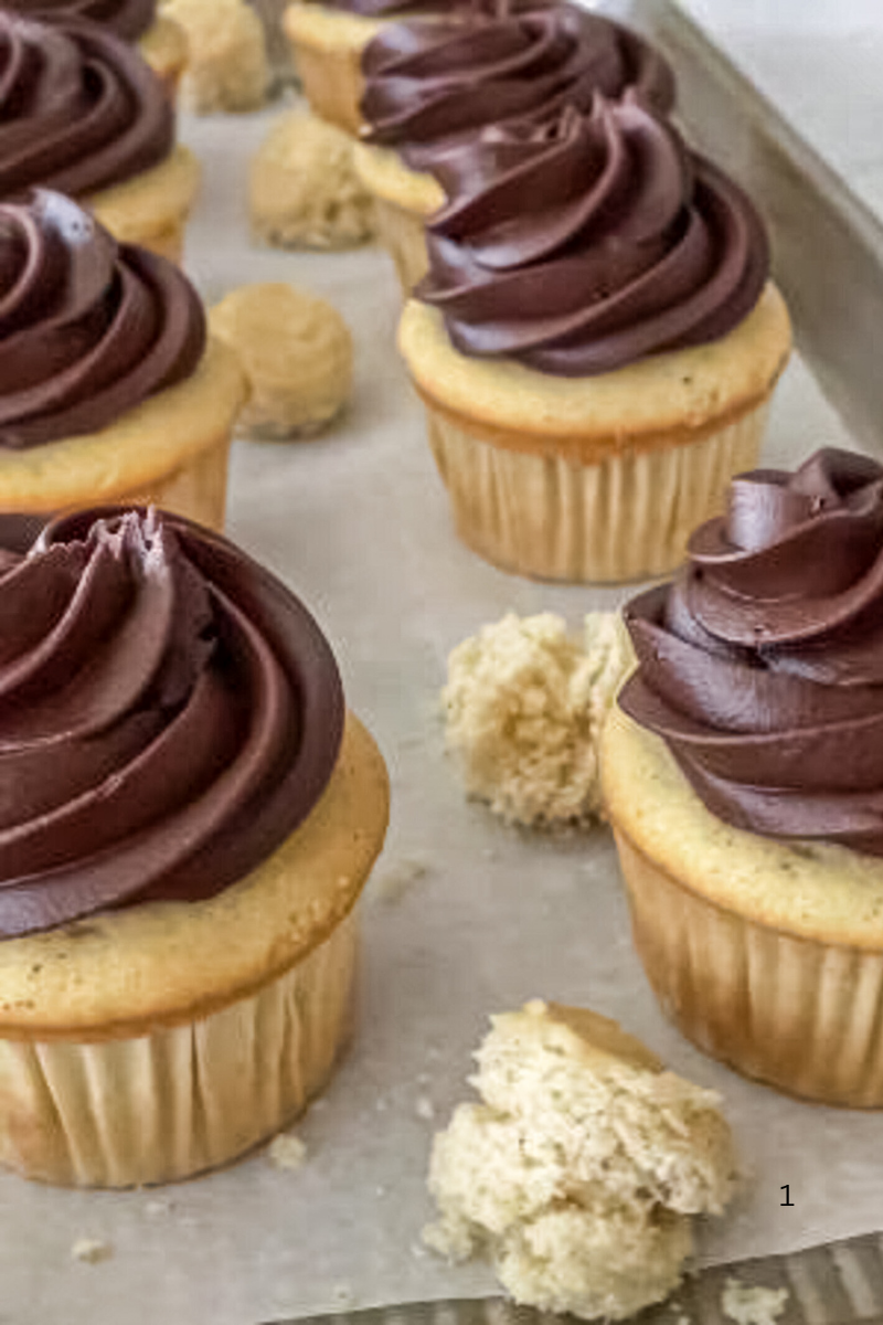 Homemade vanilla cupcakes topped with thick chocolate swirls on a parchment-lined baking sheet, with cupcake centers removed and set aside for filling.