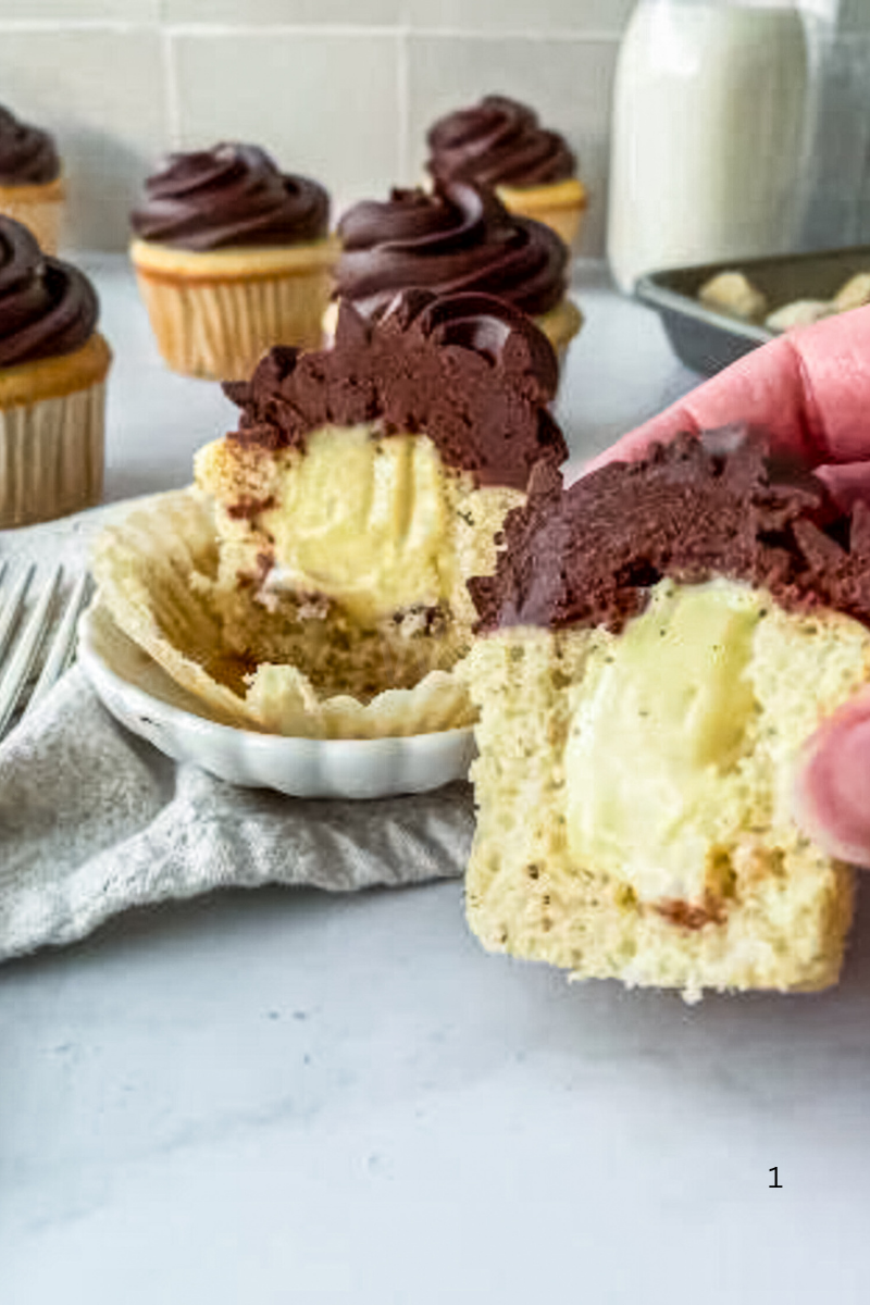 Boston Cream Cupcake cut in half to show the creamy pastry filling inside, topped with thick chocolate frosting and surrounded by more frosted cupcakes in the background.