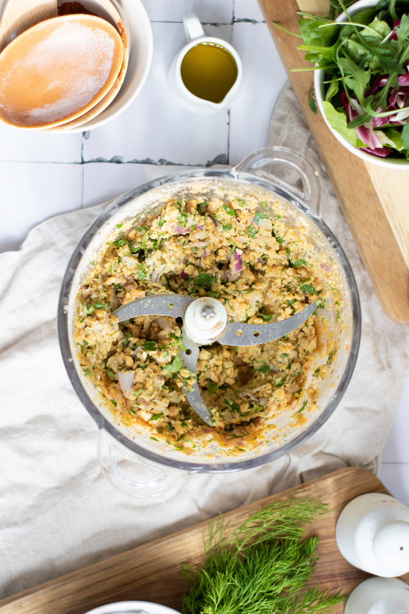Chunky falafel mixture in a food processor with chickpeas, herbs, onion, garlic, and spices ready to chill before shaping.