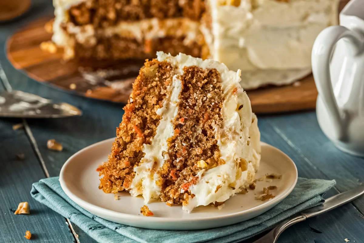 Slice of carrot cake with cream cheese frosting on a plate, with a layered carrot cake in the background