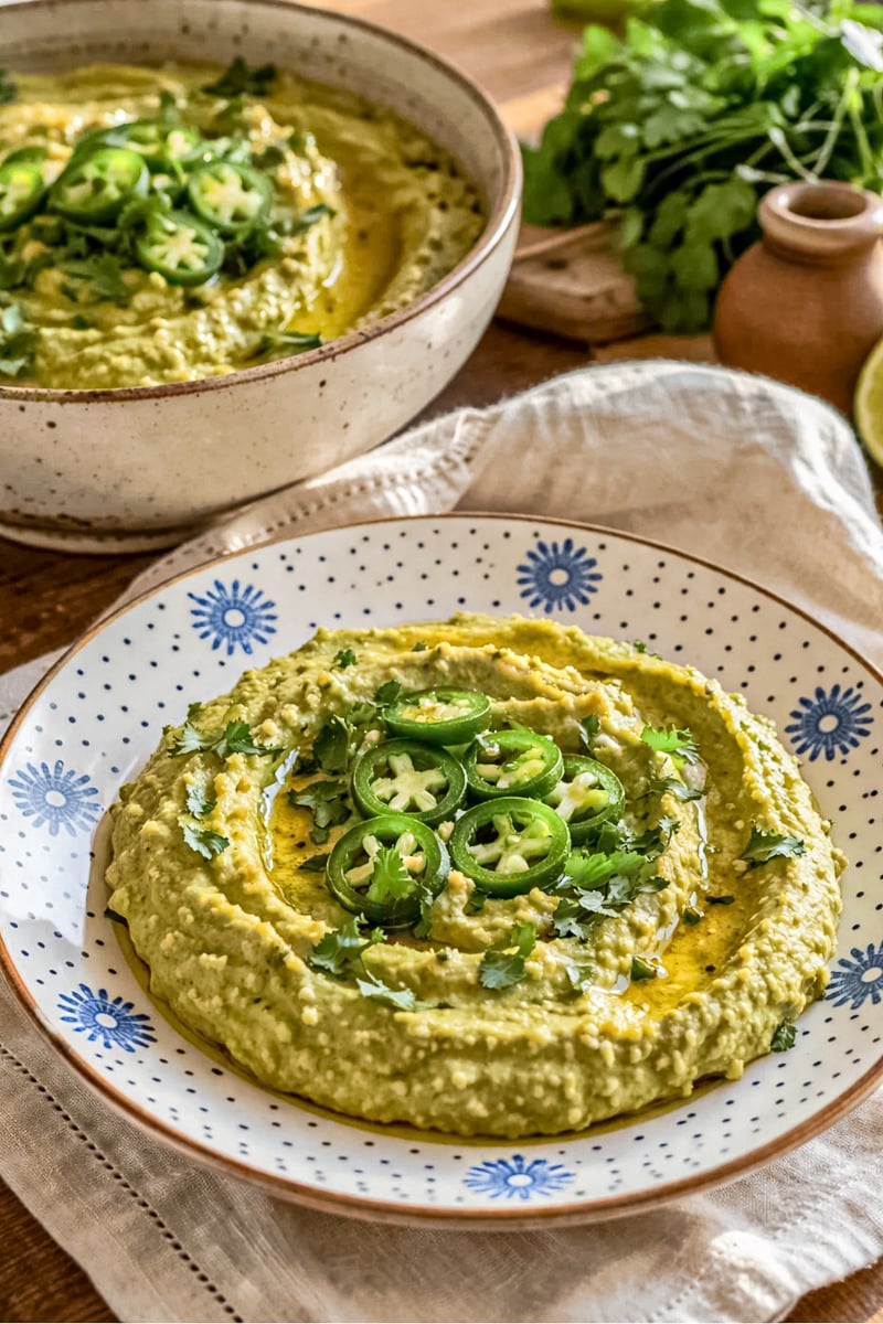 Creamy jalapeño hummus garnished with sliced fresh jalapeños, chopped cilantro, and olive oil, served in a patterned bowl with a smooth swirled texture on a rustic table.