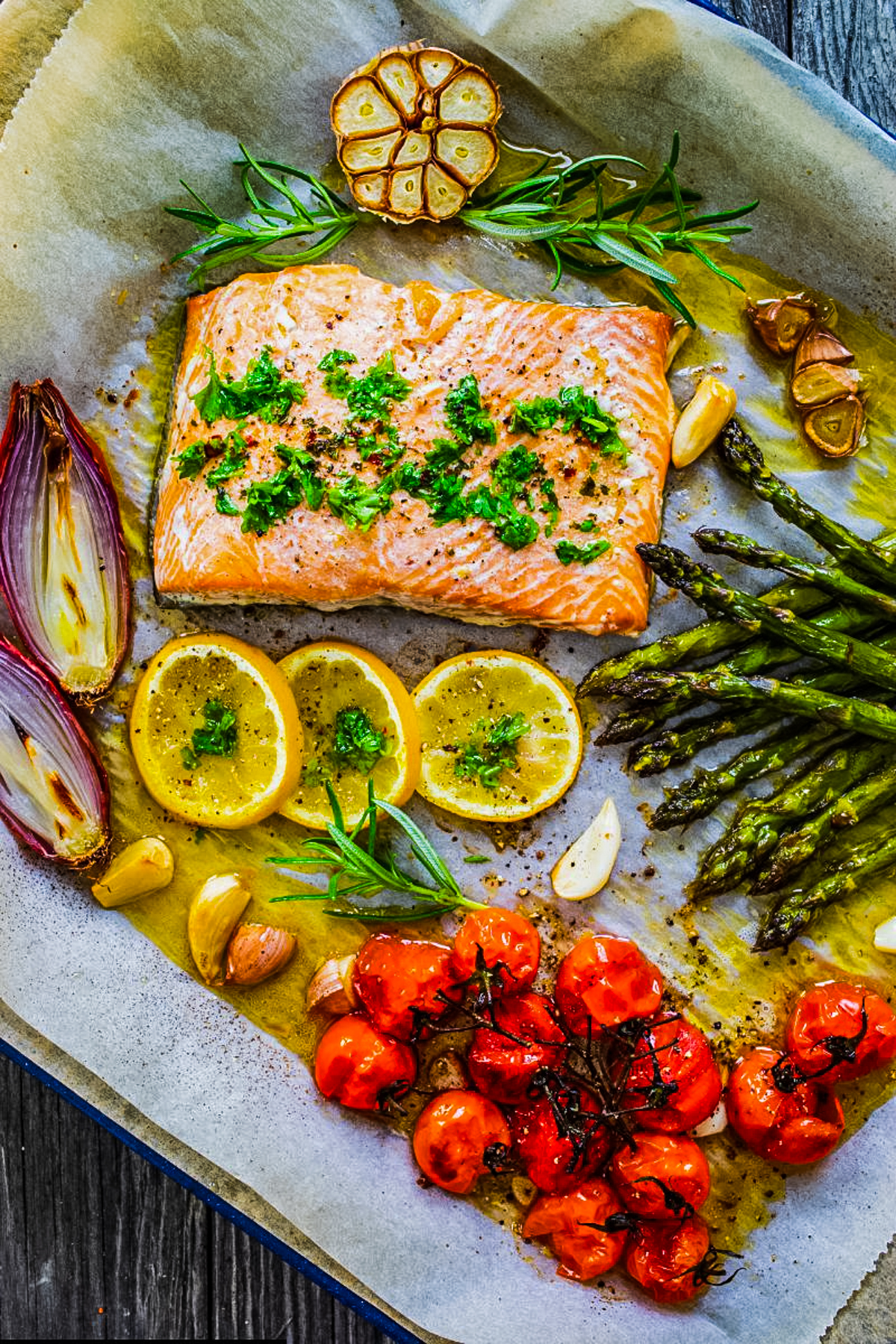 Baked salmon fillet with asparagus, lemon slices, roasted garlic, shallots, and herbs on a sheet pan for an easy high-protein dinner.
