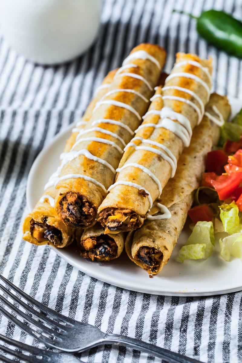 Crispy homemade chicken flautas drizzled with crema and served with shredded lettuce, diced tomatoes, lime, and jalapeño on a white plate.