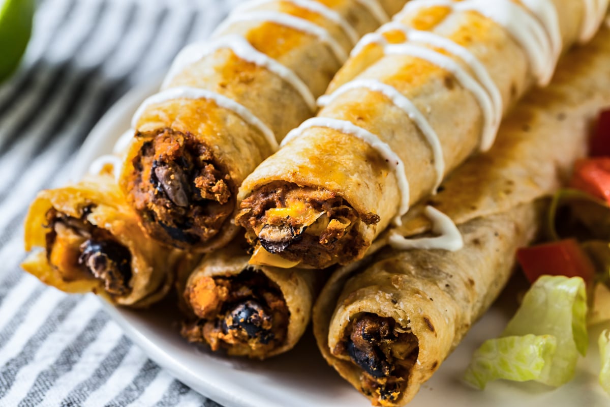 Close-up of crispy Mexican chicken flautas with seasoned chicken, black beans, and cheese filling, finished with a crema drizzle.