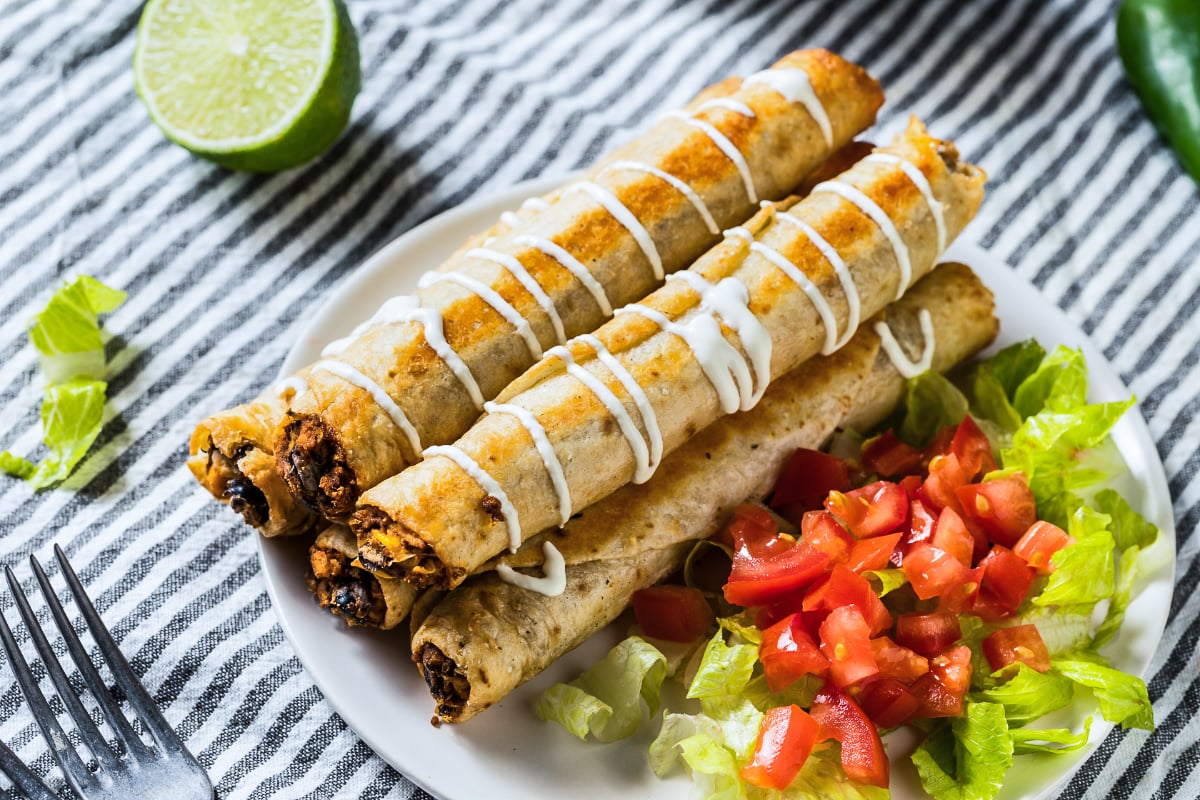 Crispy Mexican chicken flautas drizzled with crema and served on a white plate with shredded lettuce, diced tomatoes, fresh lime, and jalapeño.