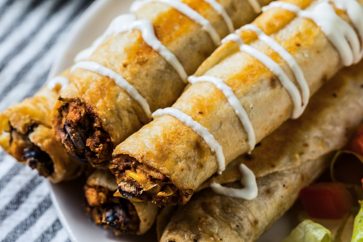 Close-up of crispy homemade chicken flautas filled with seasoned chicken and black beans, drizzled with crema and served with fresh toppings.